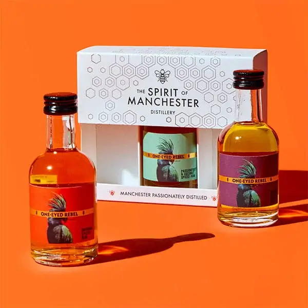 A Spirit of Manchester distillery gift set with two miniature bottles of One Eyed Rebel rum alongside a boxed bottle set, displayed against a bright orange background.